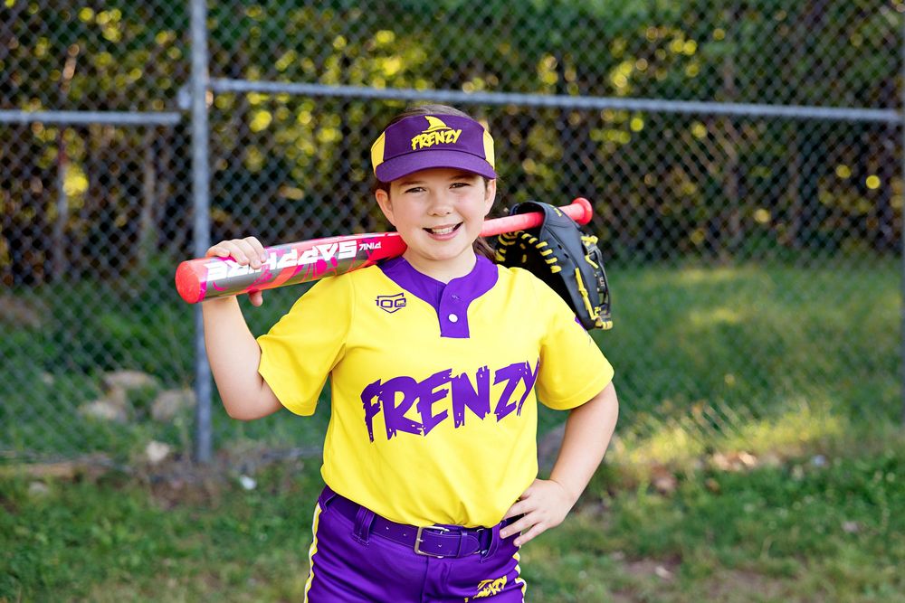 Baseball player in purple and yellow jersey holds a red bat while posing at outdoor sports field.