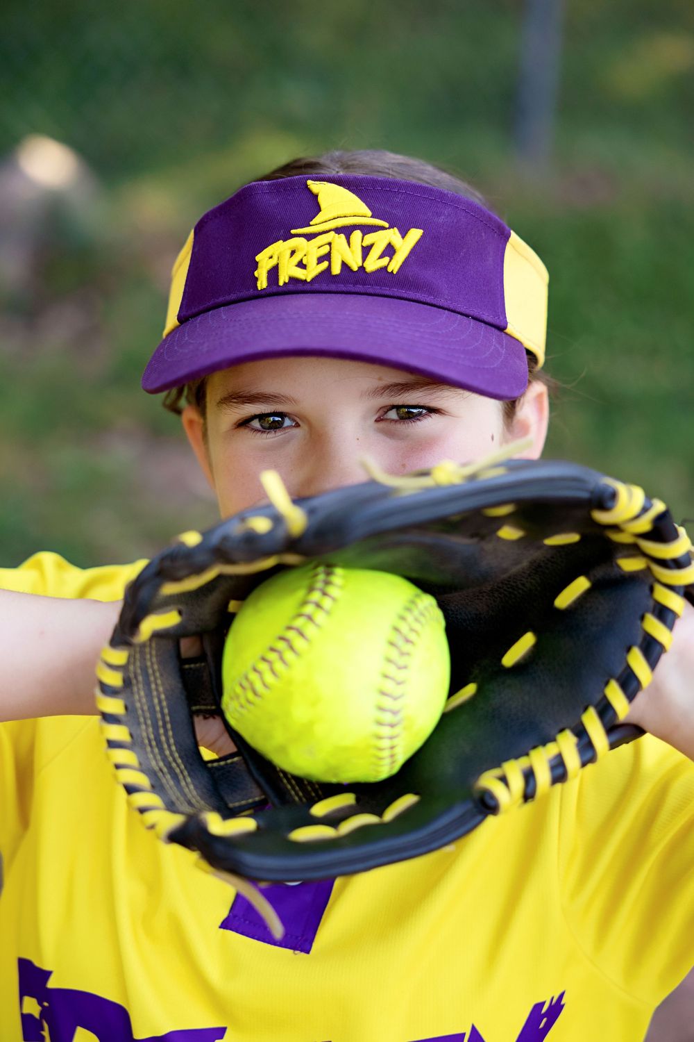 Yellow softball rests in black and yellow leather glove against purple and yellow sports uniform background.
