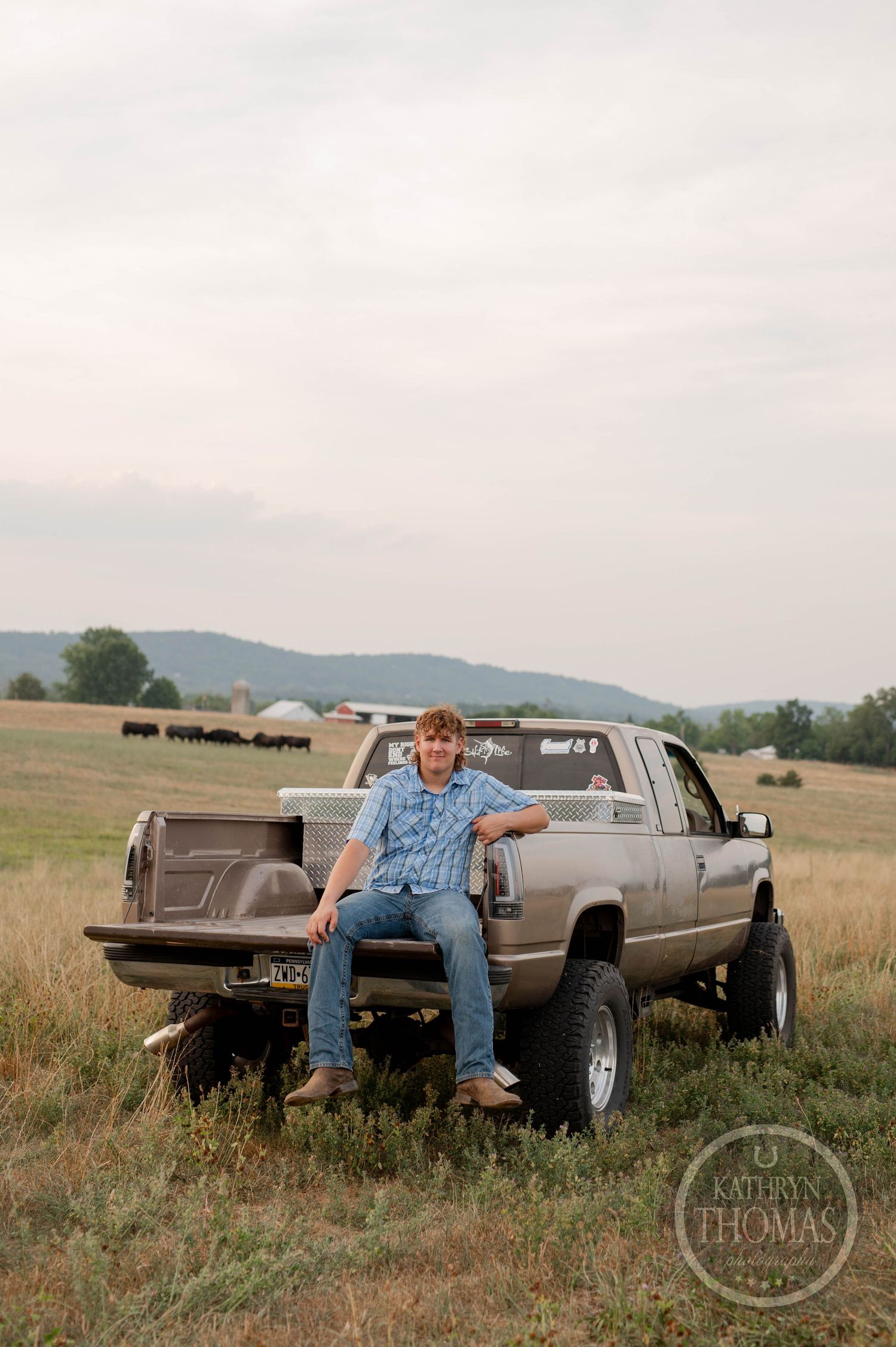 A person in jeans and plaid sits on the back of a lifted pickup truck in a grassy field.