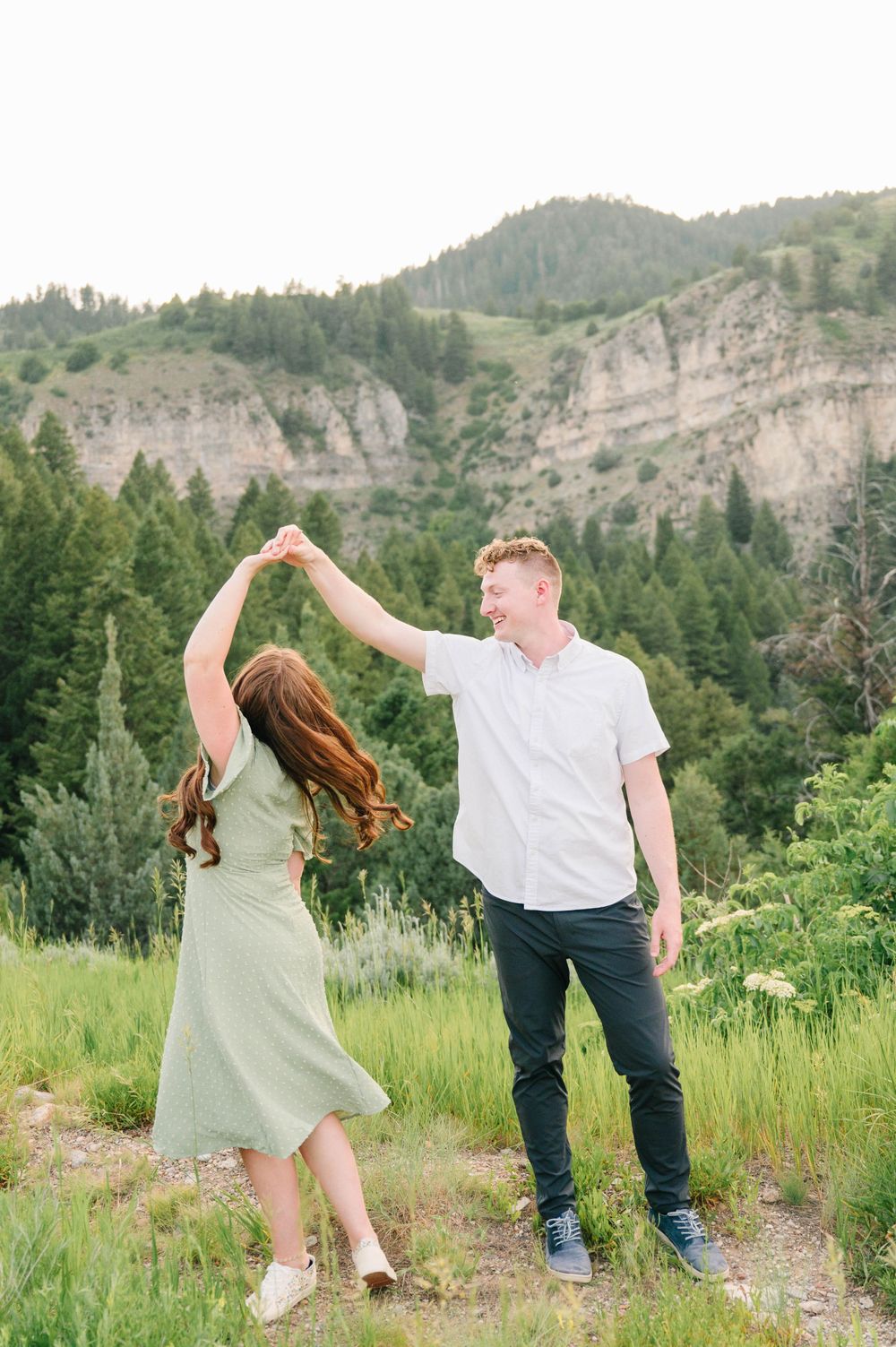 A couple dances together in a mountain meadow with dramatic cliffs and evergreens in the background.
