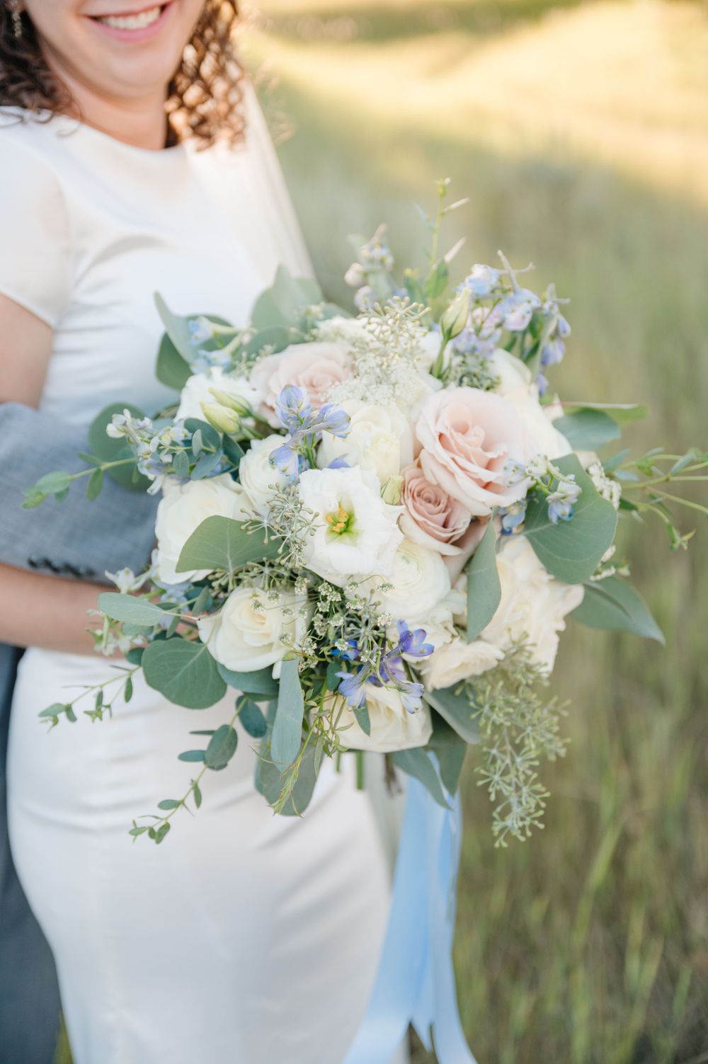 Wedding bouquet with white and blush roses, blue flowers, and cascading greenery against golden light.