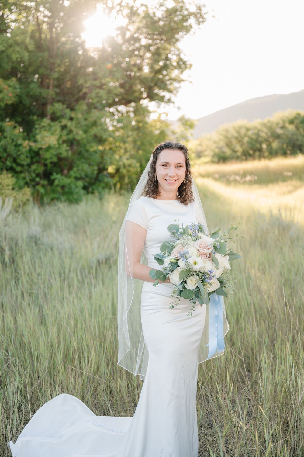 Bride in fitted white dress holds elegant bouquet while standing in sunlit mountain meadow.