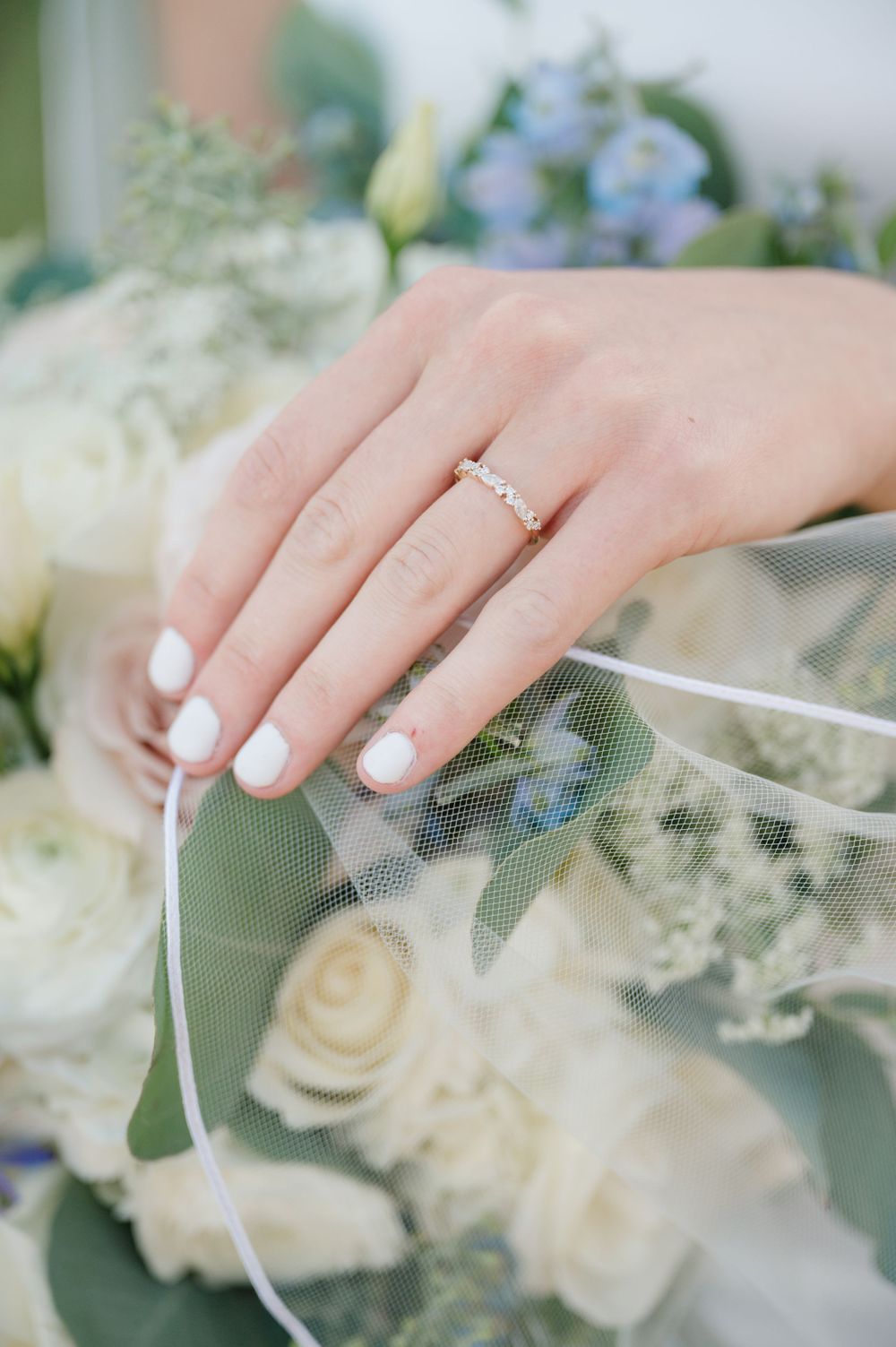 Delicate engagement ring displayed on manicured hand with white roses and blue flowers in background.