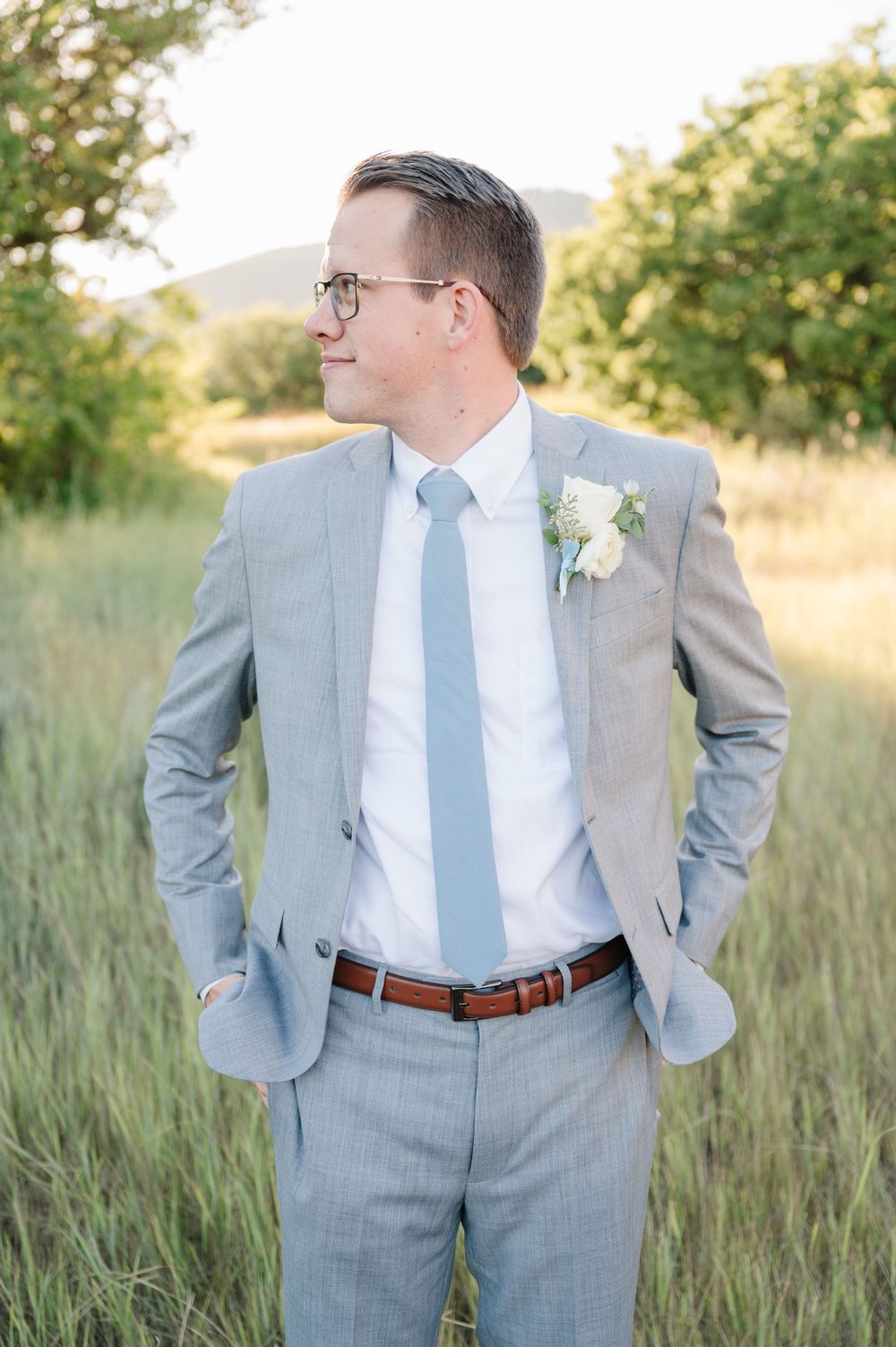 Groom in classic gray suit and light blue tie poses in sunlit mountain meadow.