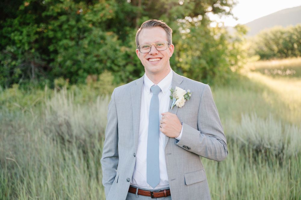 Groom in light gray suit and blue tie smiles while standing in sunny mountain meadow.