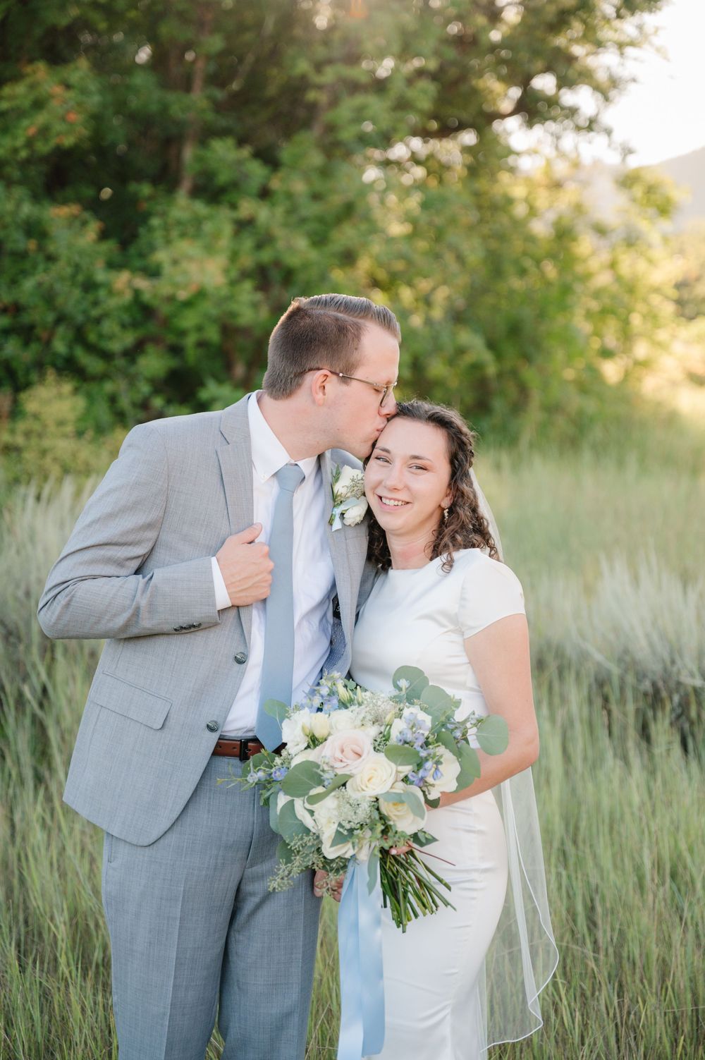 Tender wedding moment captured in tall grass with bride holding delicate blush and blue bouquet.