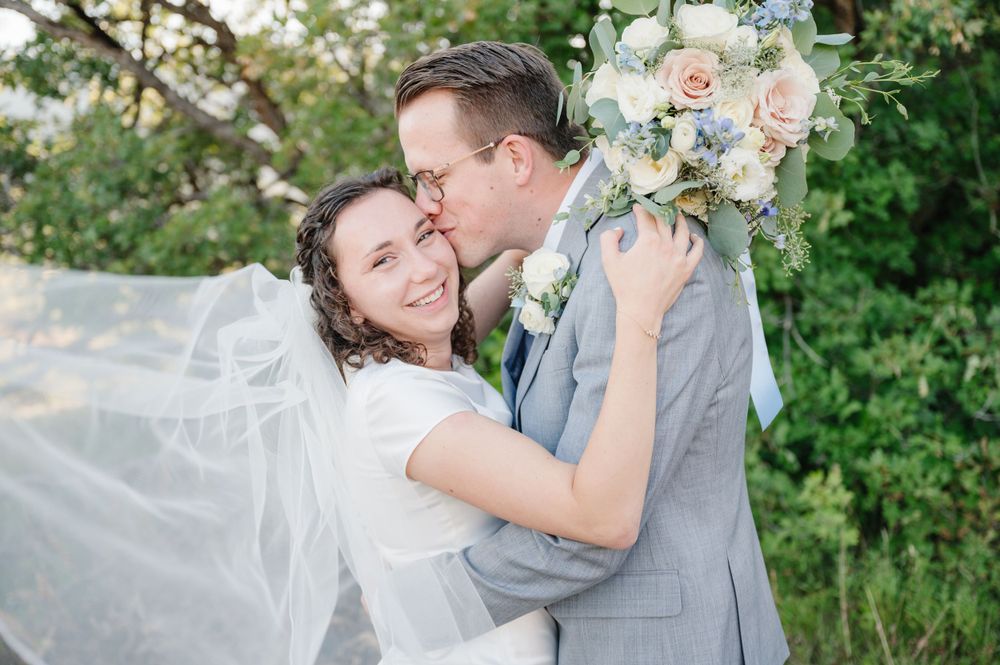 Romantic wedding embrace with flowing veil and pastel bouquet against green backdrop.