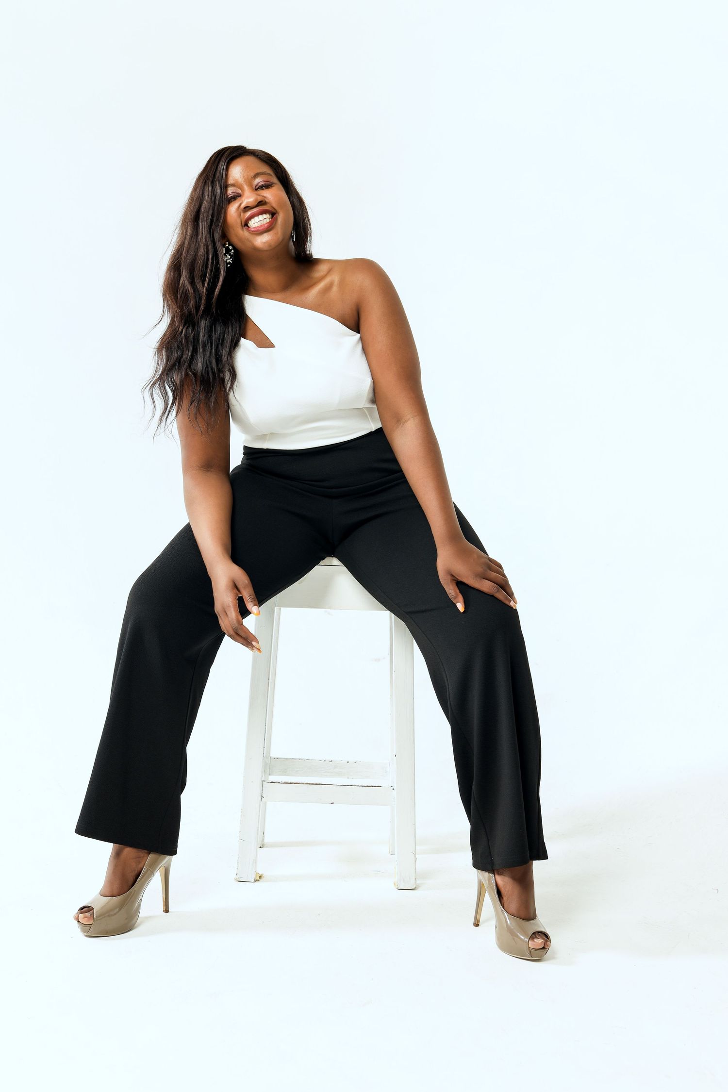 Woman in black pants and white top posing playfully on white chair against light background.