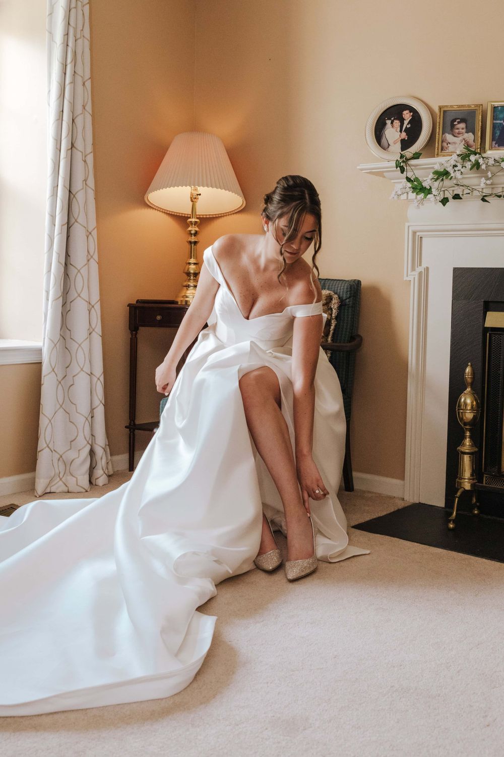 Elegant wedding dress preparation scene with table lamp, patterned curtains and fireplace in warm-lit bedroom.