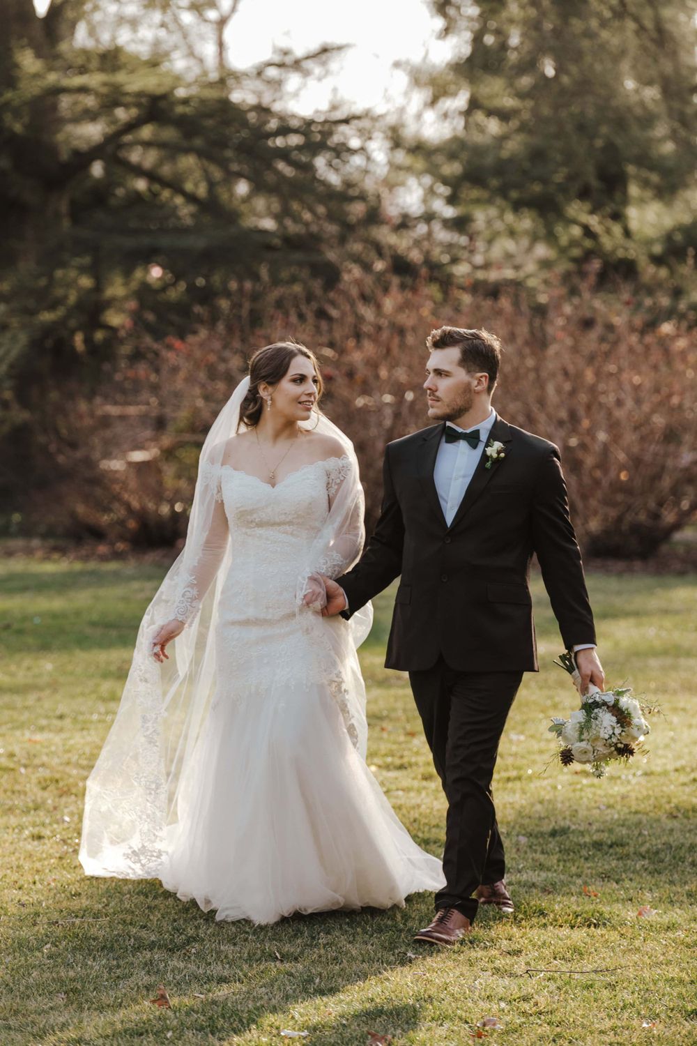 A romantic wedding couple walking together on grass with greenery in the background during a fall or winter day.