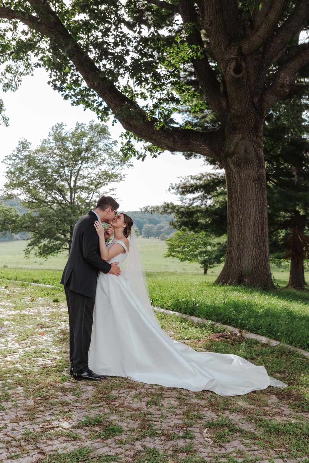 A couple shares their first kiss as newlyweds under a large oak tree in a scenic countryside setting.