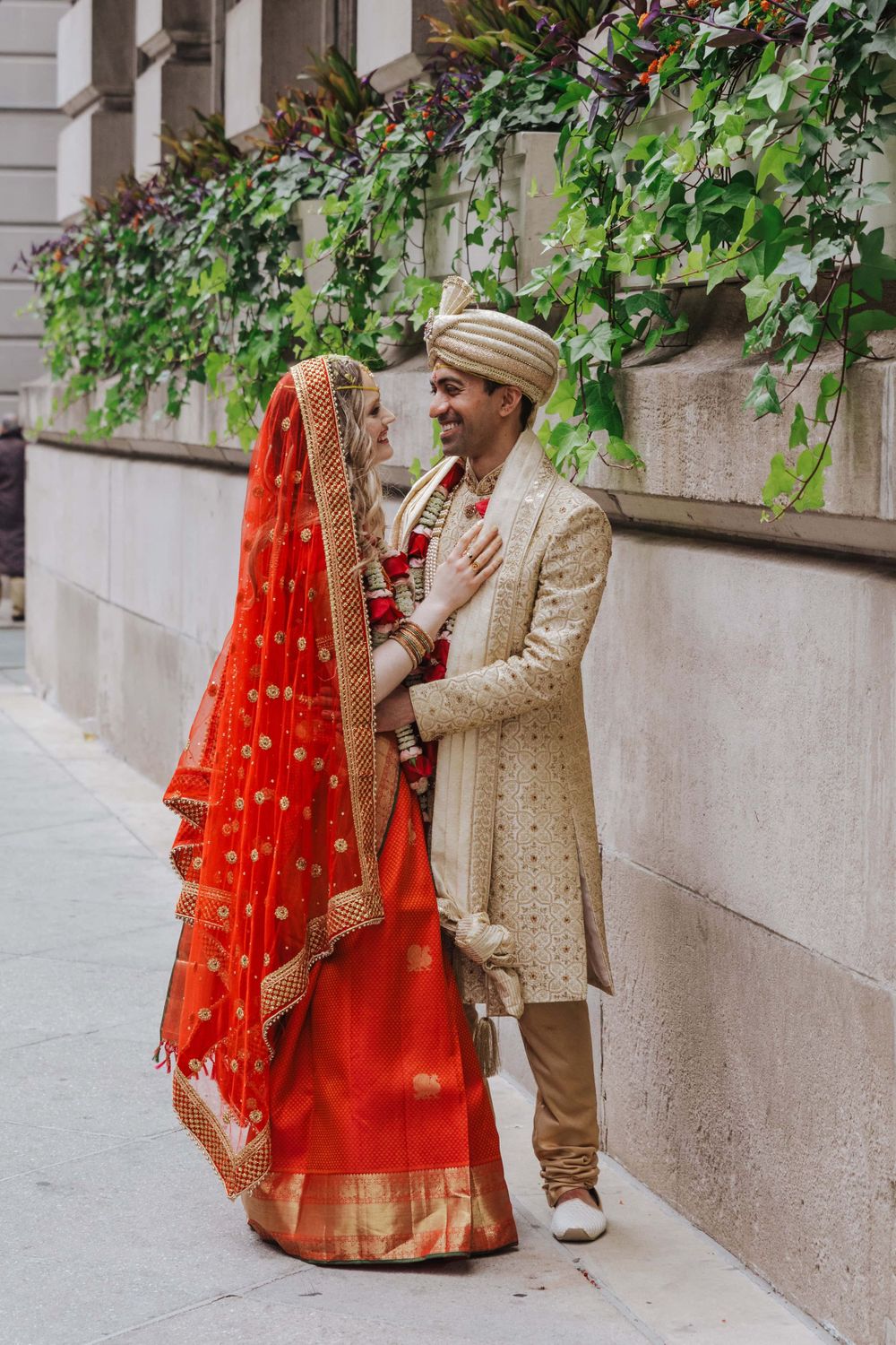 Traditional Indian wedding attire featuring a bright red sari with gold embellishments and cream sherwani against stone wall.