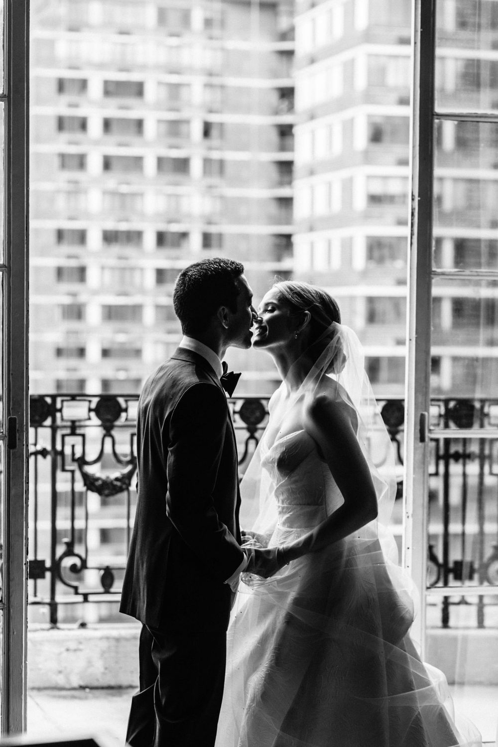A romantic black and white wedding photograph showing a couple in silhouette kissing against a city backdrop.