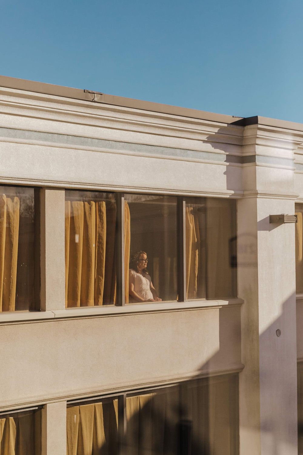 A sun-lit apartment building exterior with beige walls and gold curtains visible through a window on the upper floor.