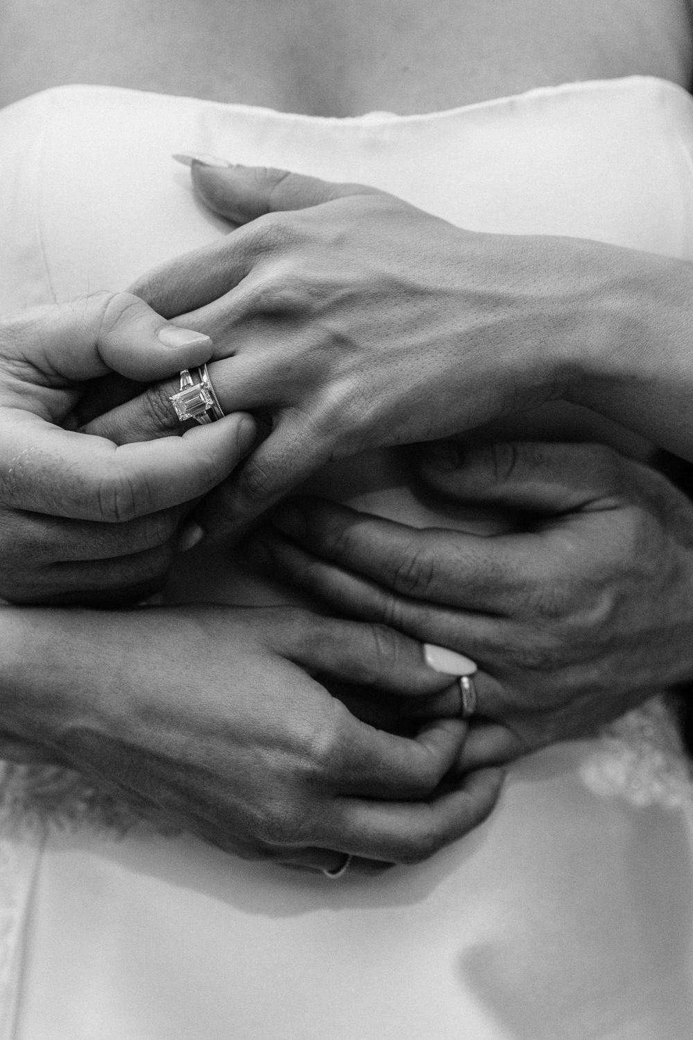 Black and white close-up of hands wearing wedding rings against white fabric showing an intimate moment.