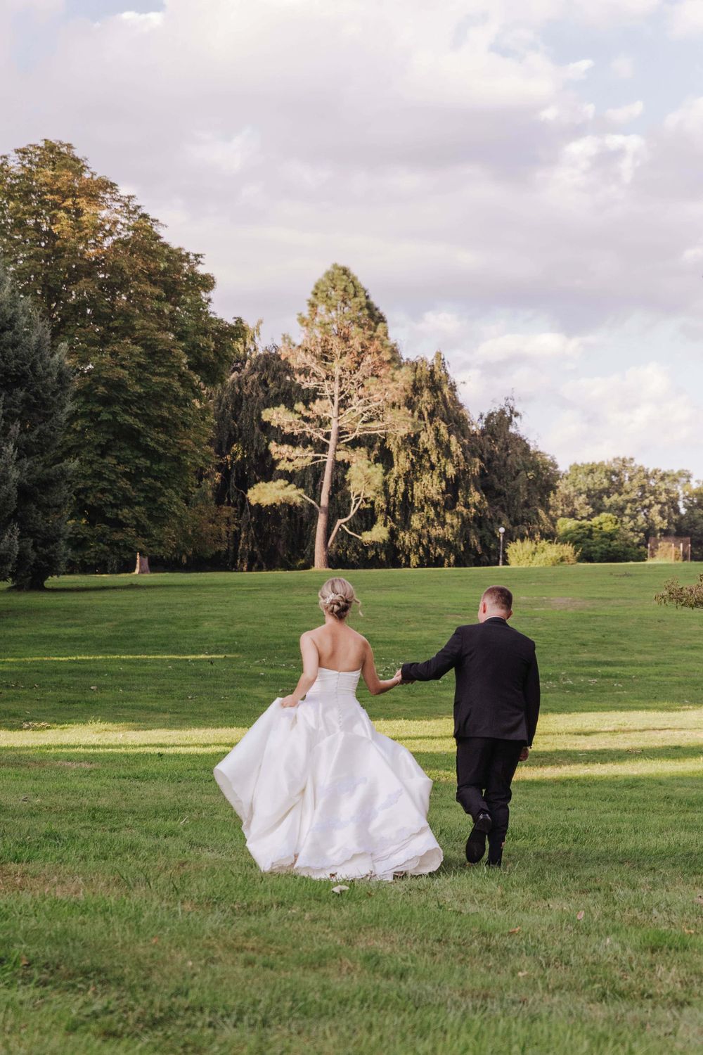 Newlyweds walk hand in hand across a lush green lawn with tall trees in the background.