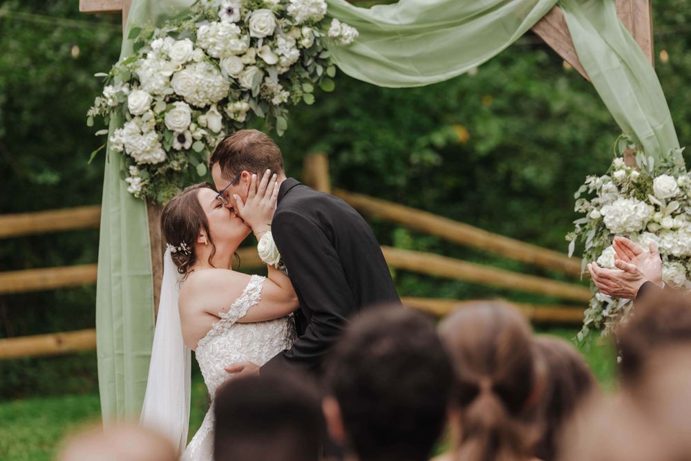 A romantic wedding ceremony moment under a floral arch draped with white fabric and roses in an outdoor garden setting.