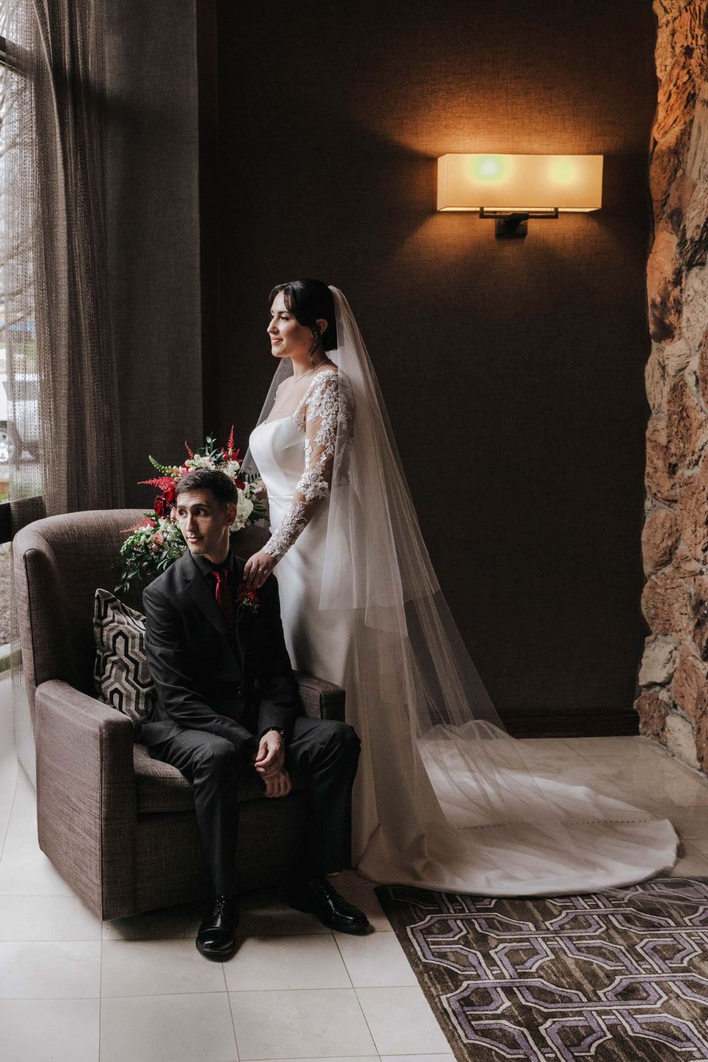 An elegant wedding photograph featuring a white lace gown with flowing veil in a warmly lit indoor setting with stone walls.