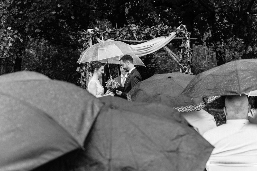 A wedding couples veil blows in the wind above a sea of umbrellas during a rainy outdoor ceremony.