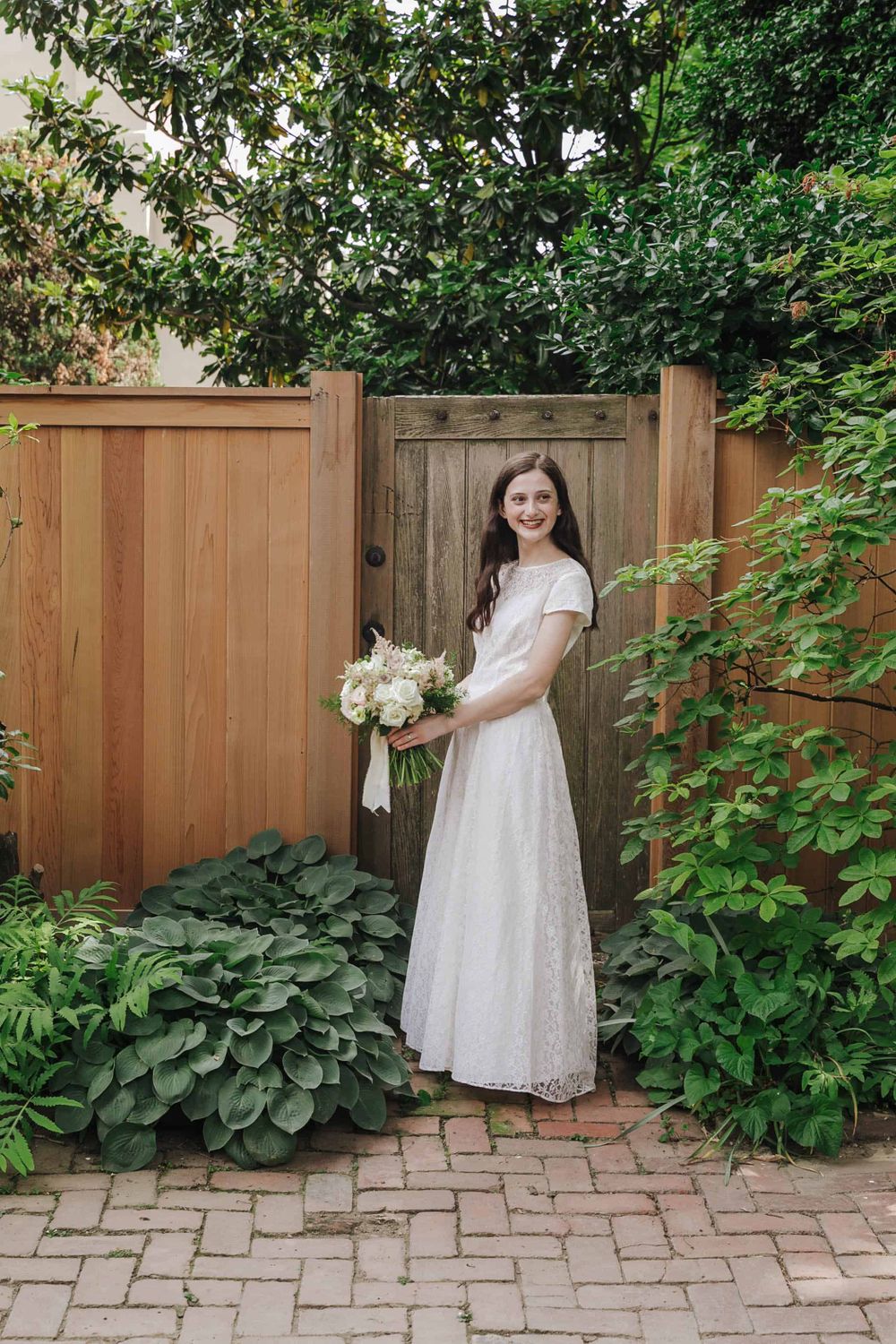 A person in a white lace dress stands in a garden setting with a bouquet by a wooden fence and gate.