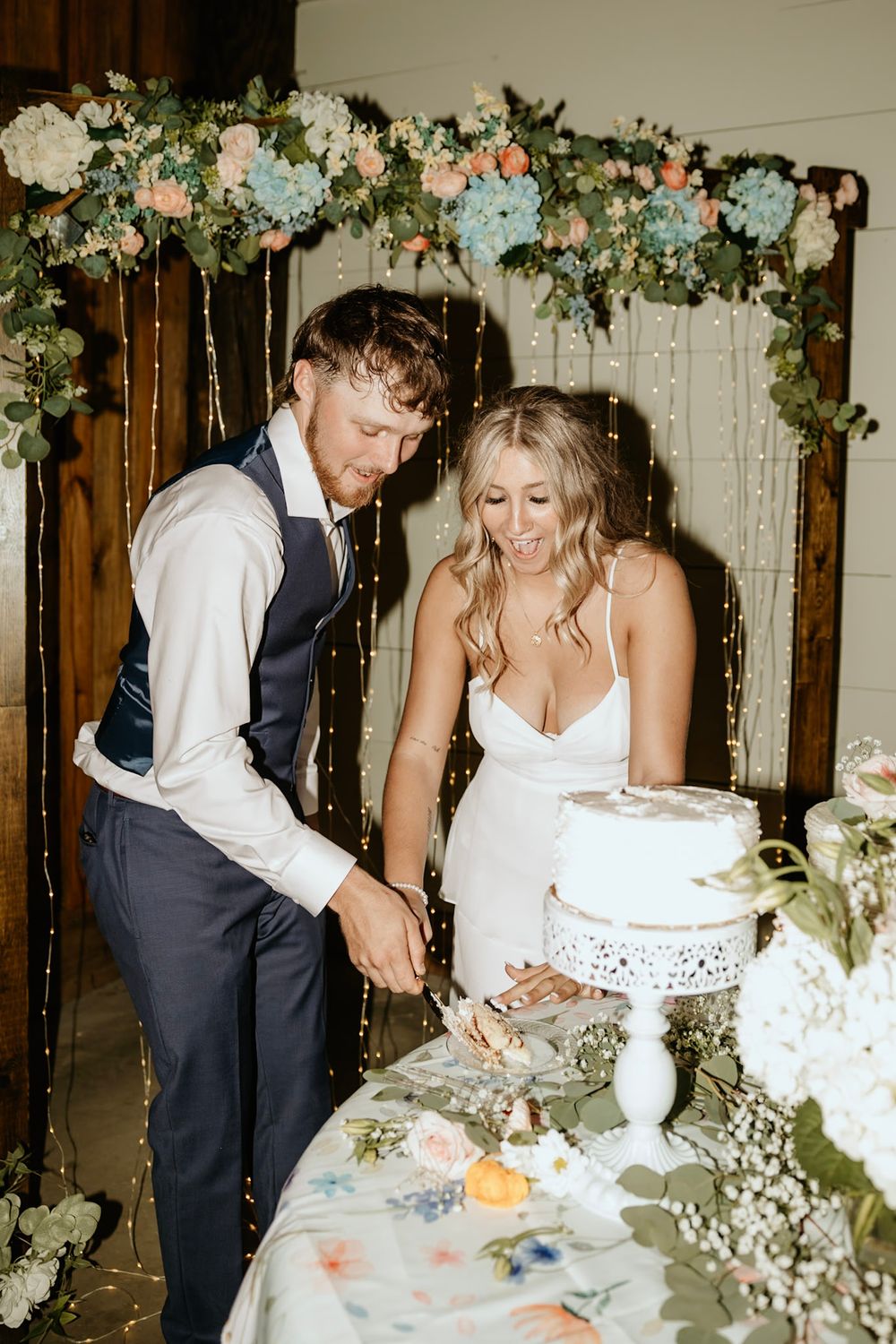 A couple cuts into their elegant white wedding cake beneath a floral arch decorated with pale blue and peach blooms.
