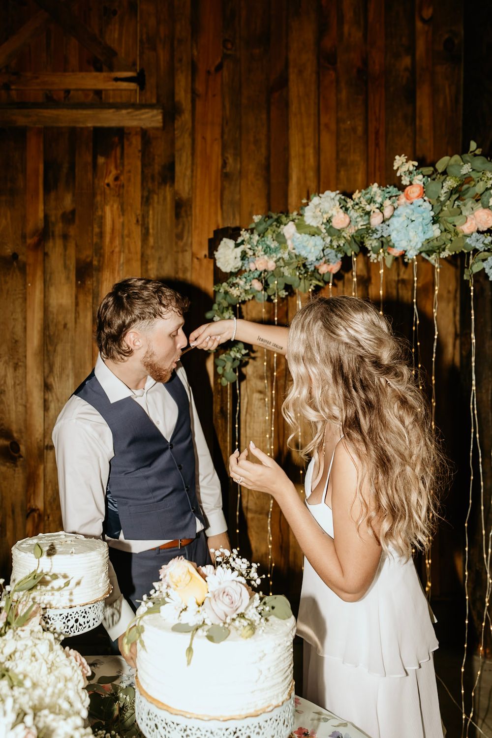 A couple shares cake at their rustic barn wedding reception with floral decor and string lights in the background.