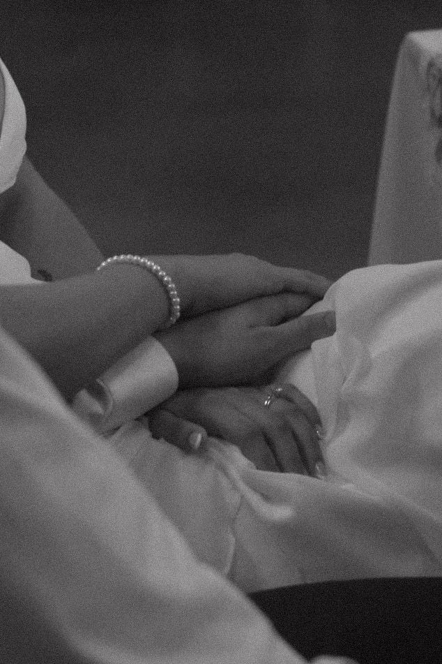 A black and white closeup of clasped hands showing a delicate pearl bracelet against white fabric.