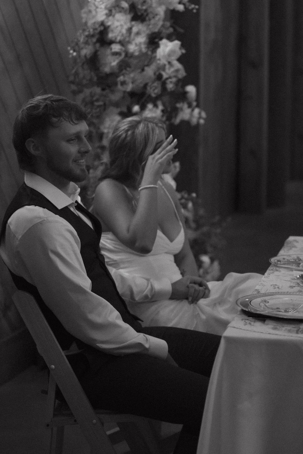 A black and white wedding photo showing a couple seated at a reception table with floral decorations.
