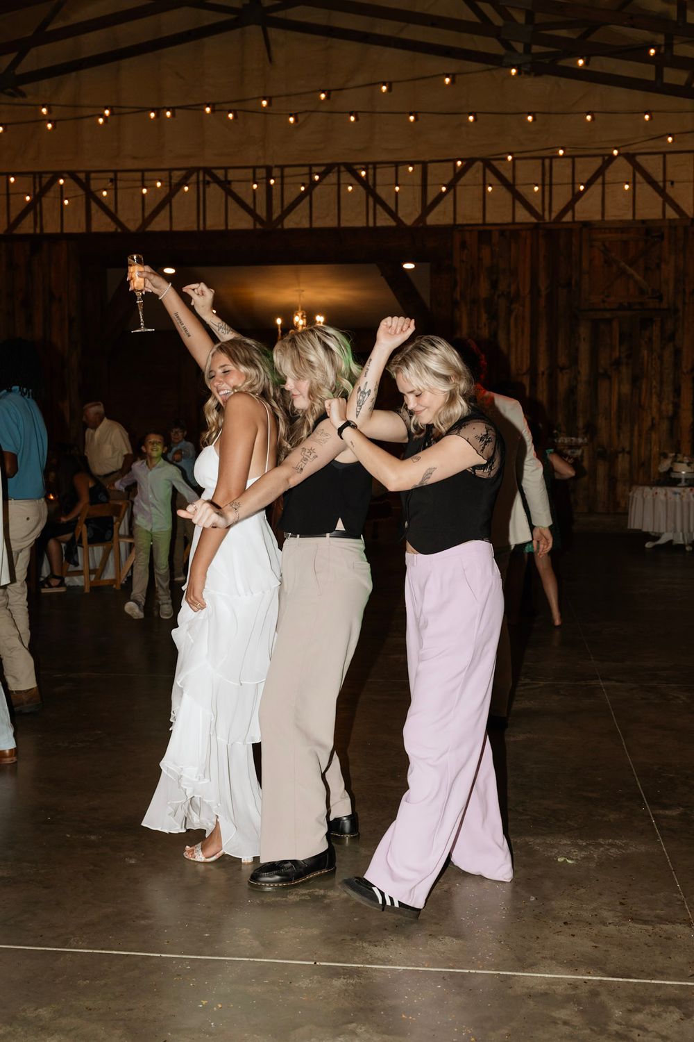 A group of party guests in elegant dresses dancing together with raised arms under string lights at a rustic venue.