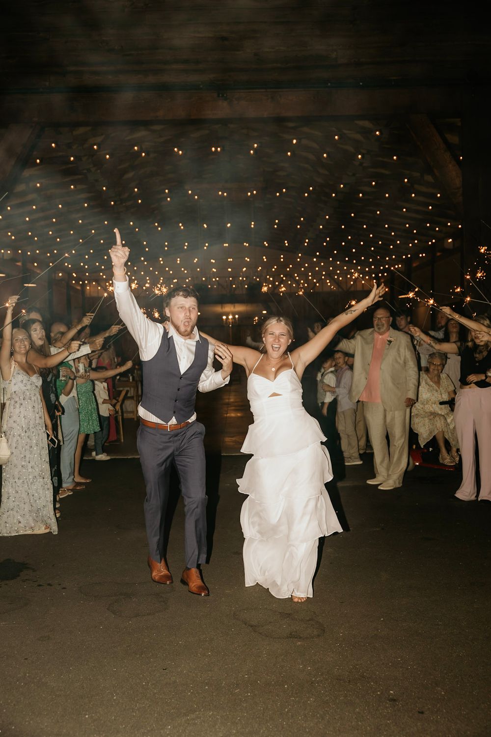 Wedding celebration under twinkle lights with couple dancing joyfully at their rustic barn reception.
