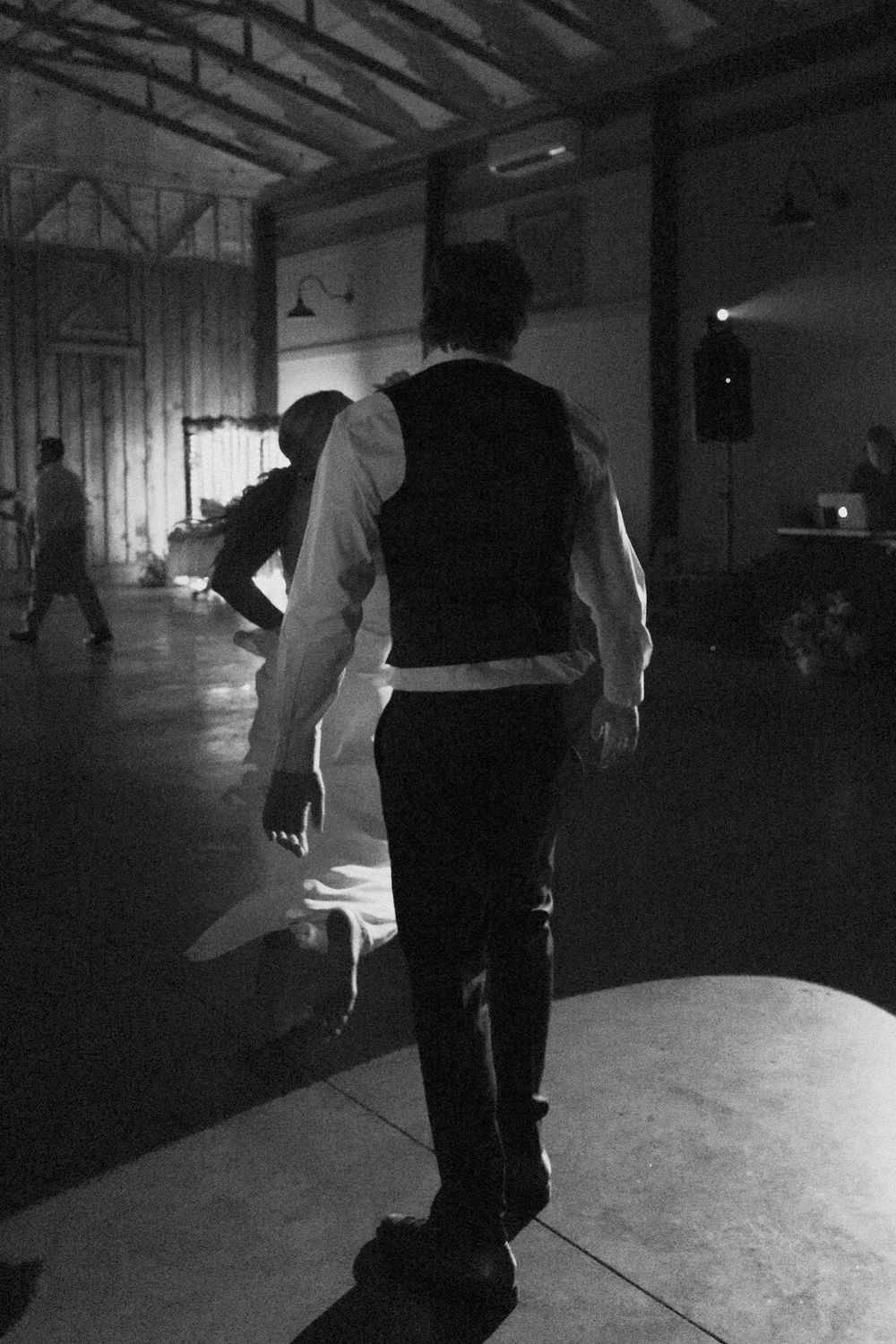A black and white photo shows someone in formal attire and a vest walking across a dimly lit dance floor or stage.
