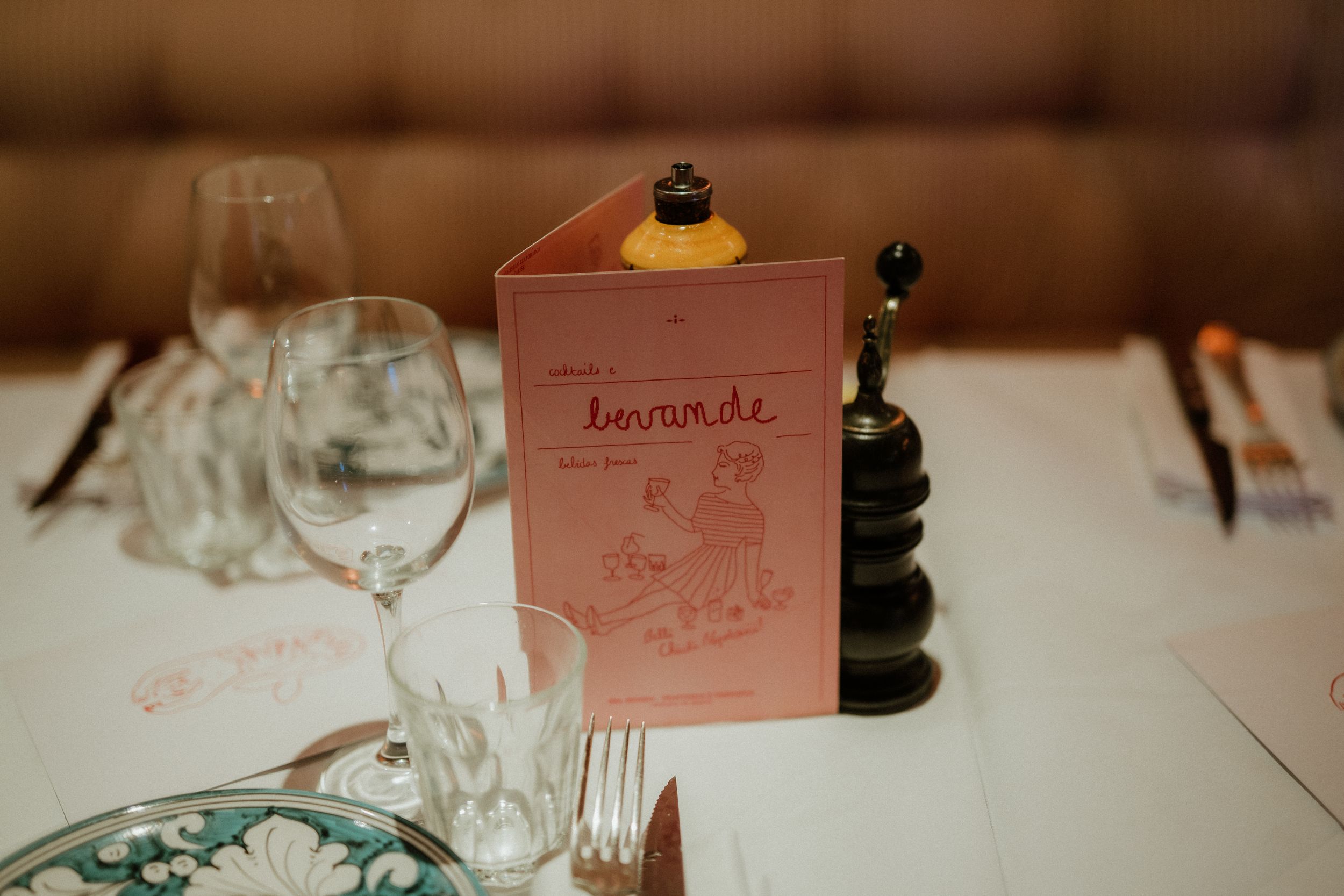 Pink menu card stands on white table setting with empty wine glasses and black pepper grinder at restaurant.
