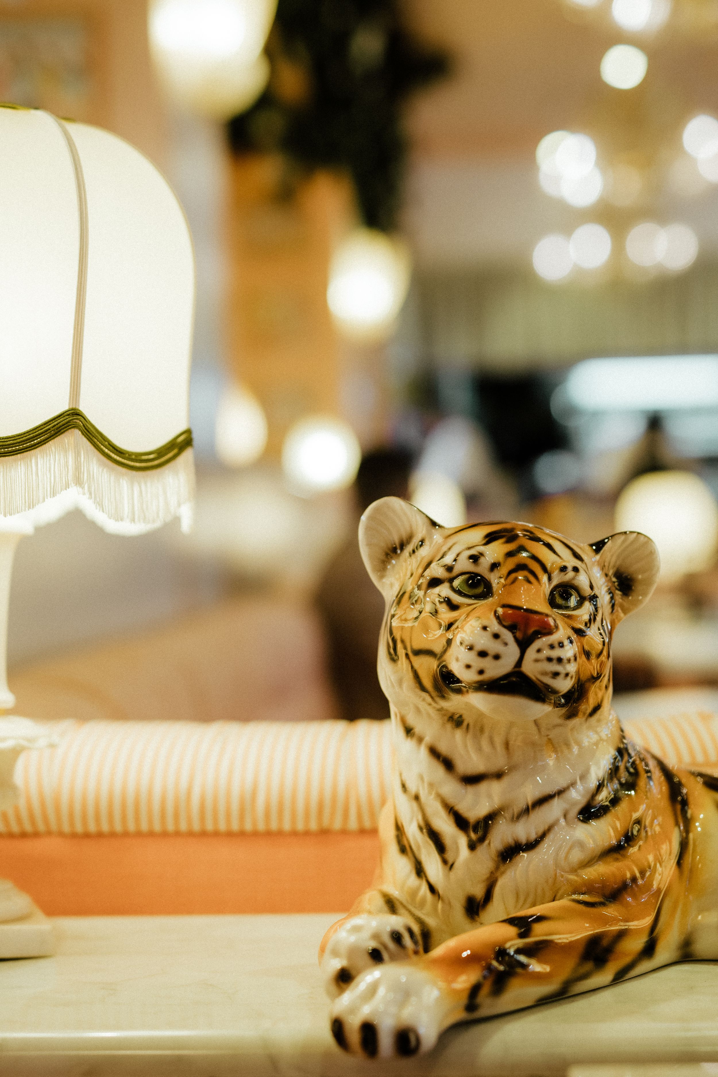 Decorative ceramic tiger figurine sitting on a table with blurred lamp and bokeh lights in the background.
