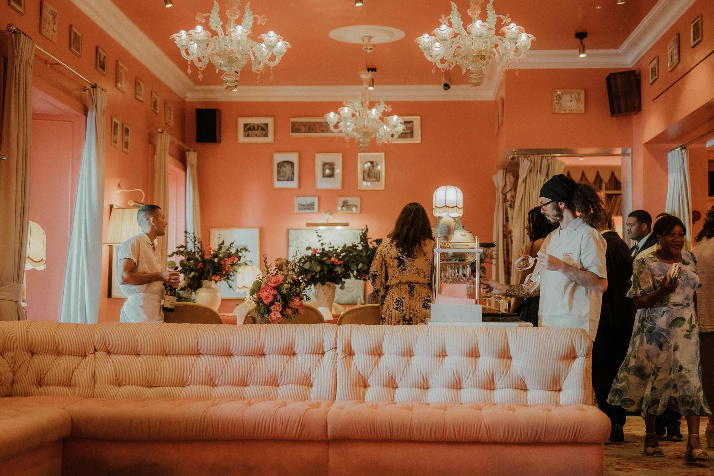Elegant salmon-colored room with crystal chandeliers, tufted seating, and floral arrangements during a special event.