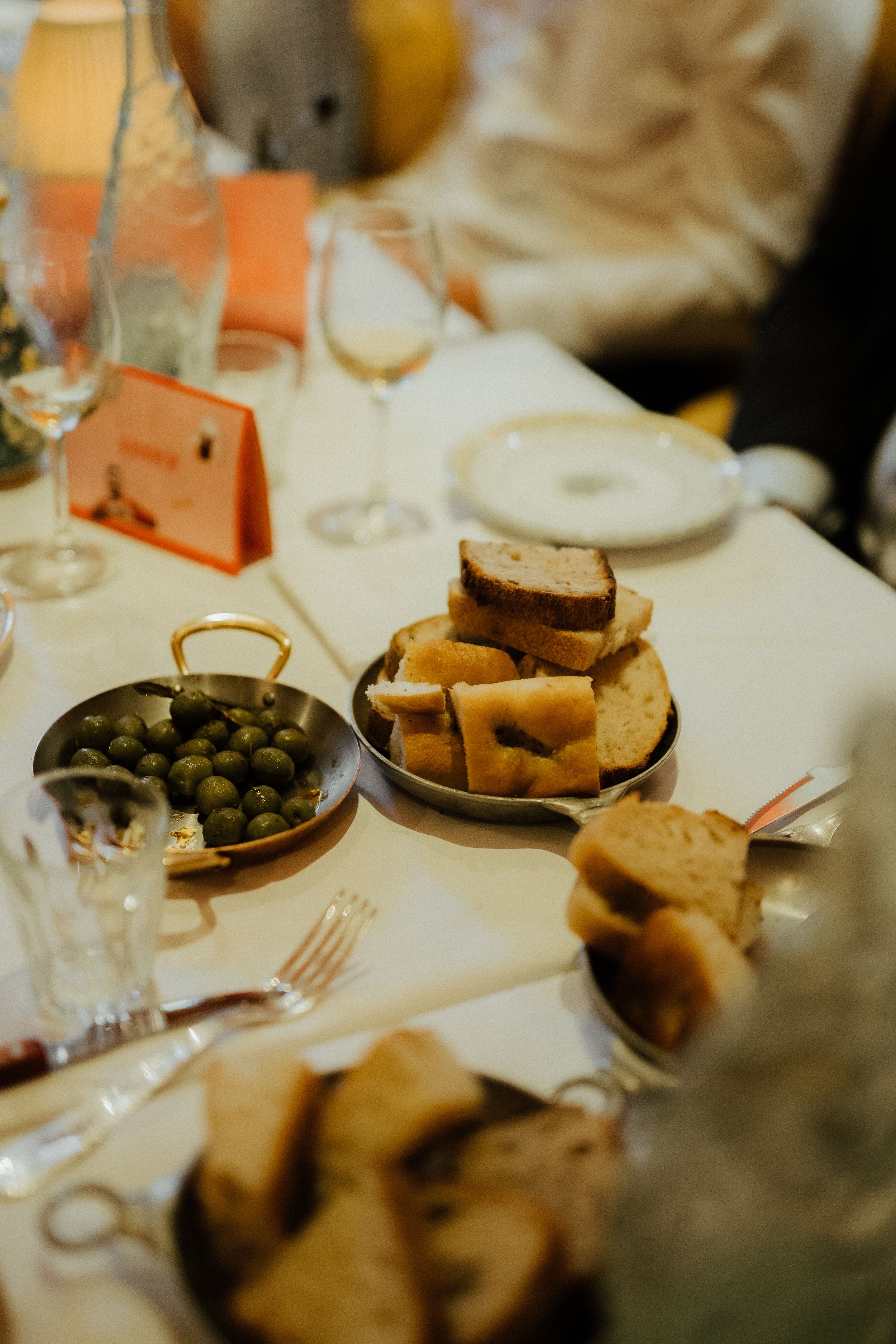 Close-up view of an elegant dinner table setting with bread, olives, and wine glasses on white tablecloth.