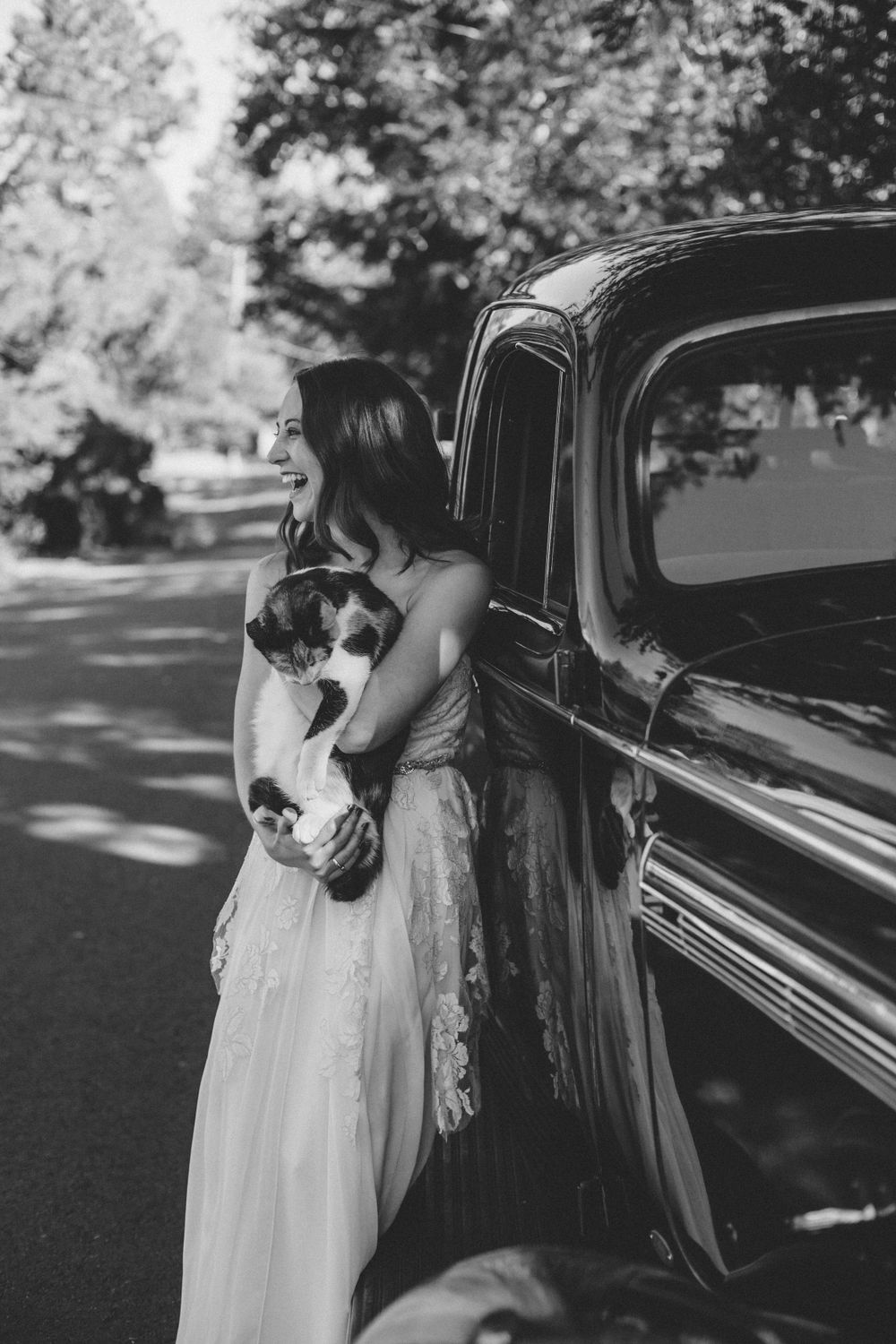Bride standing beside a vintage car, holding her cat, captured in a timeless black-and-white wedding portrait. St Helena, CA
