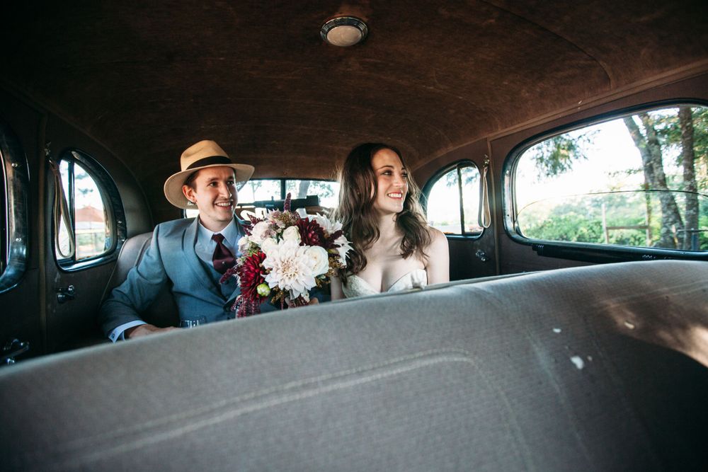 Newlyweds sitting in the back of a vintage car, holding a bouquet, with soft natural light streaming through the windows. St Helena, CA