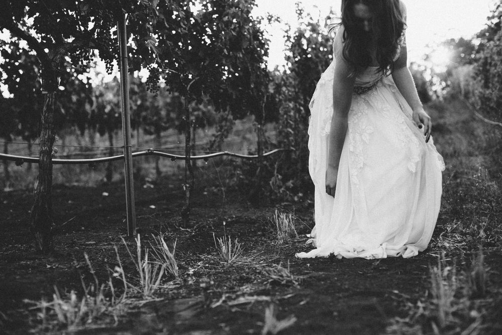 Bride in a flowing dress, sitting on a stone wall in a vineyard, captured in an elegant black-and-white moment. St Helena, CA