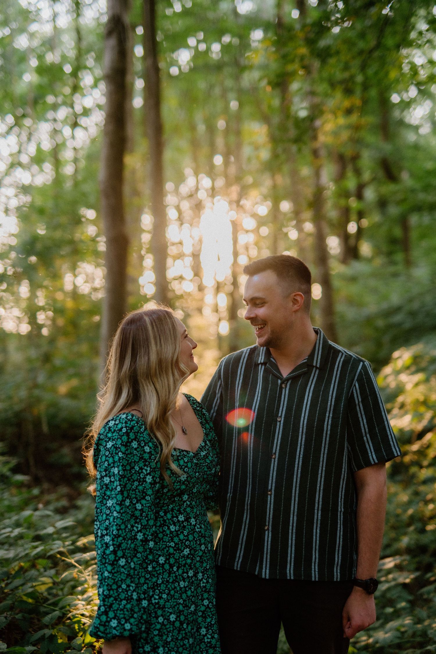 A couple shares a joyful moment together in a sunlit forest, wearing green floral dress and striped shirt.
