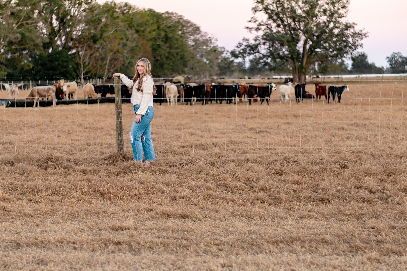 A high school senior stands in a dry pasture with a large herd of cattle grazing in the background.