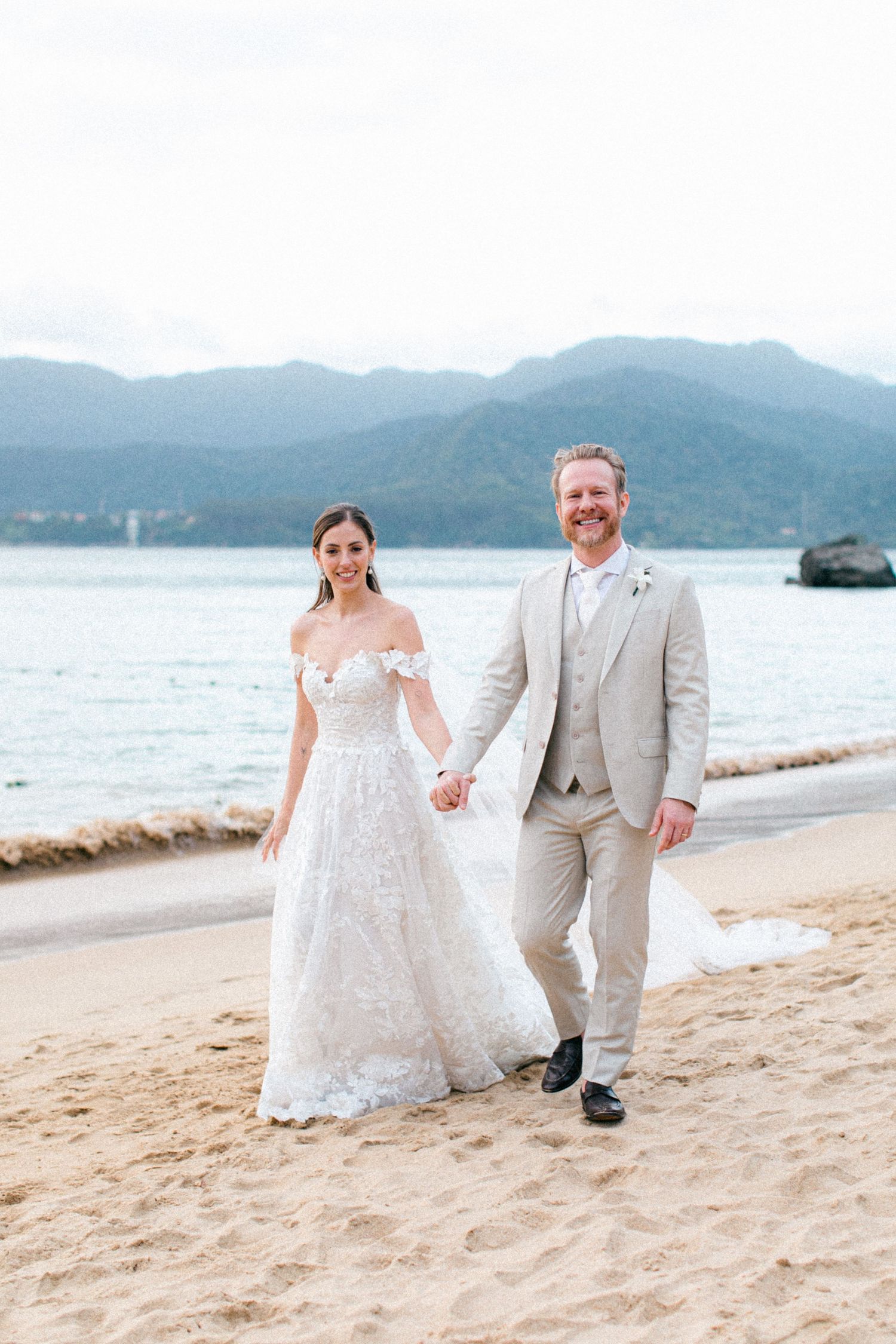 Newlyweds walking hand in hand on a scenic beach with mountains and water in the background.