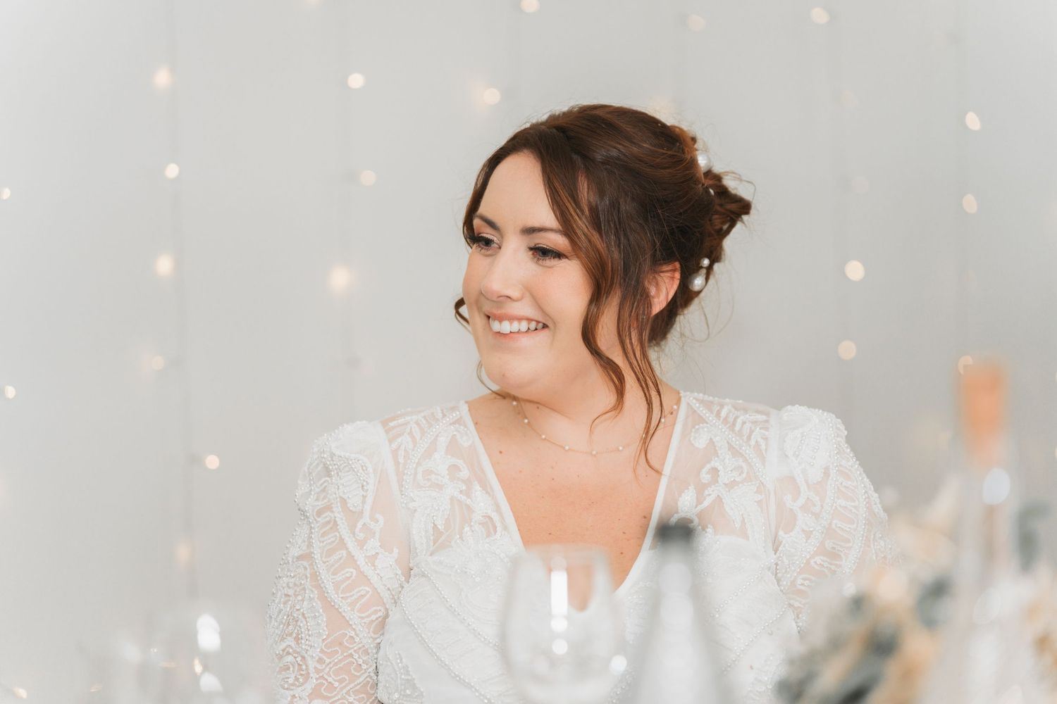 A smiling bride wearing a white lace dress poses amidst glittering bokeh lights.