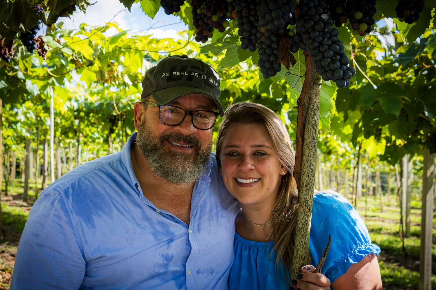 Portrait of smiling couple in vineyard under green vine canopy, photographed by Piret Victoria Ribas.