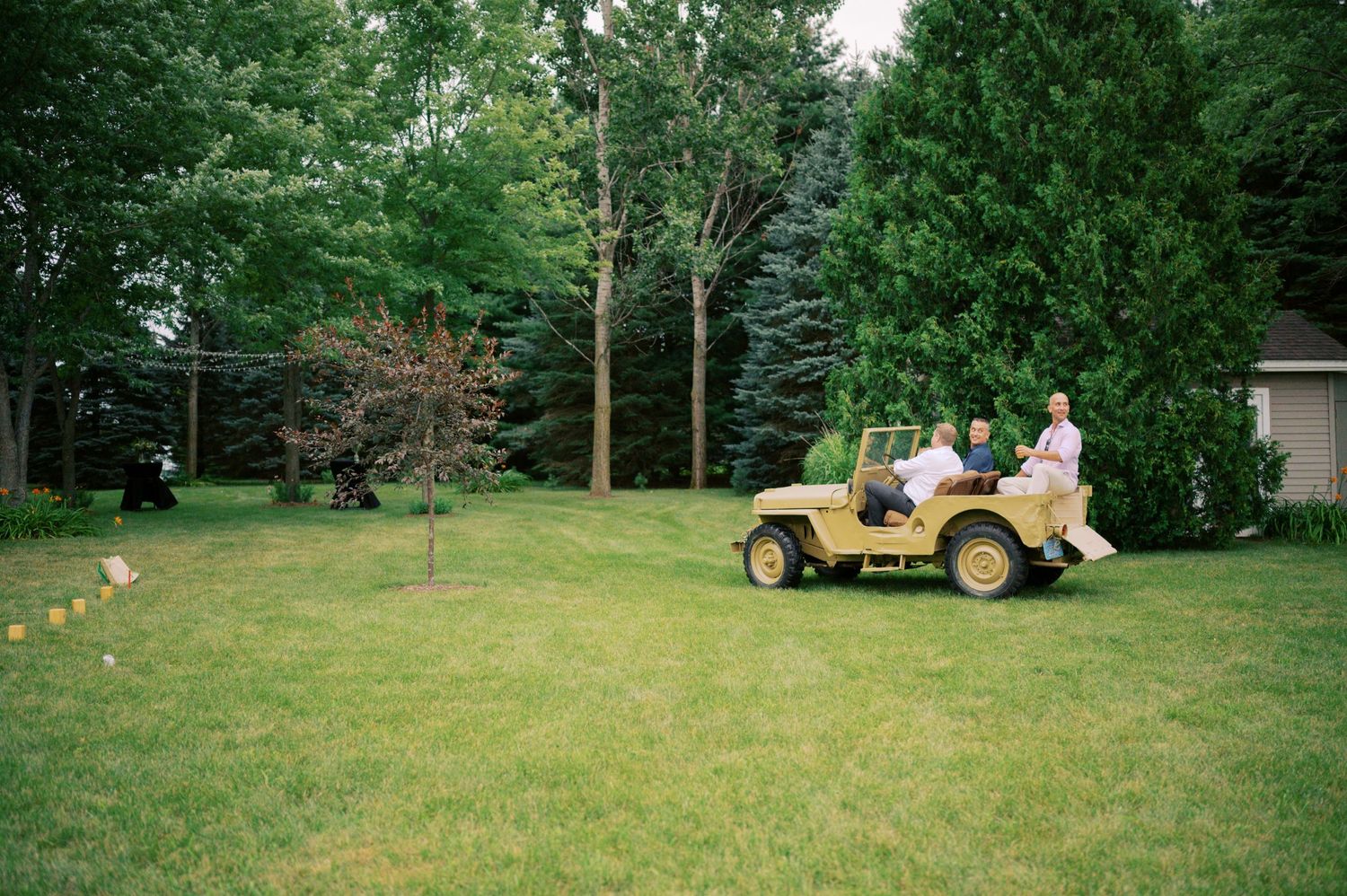 Two people on a yellow utility vehicle on a grassy lawn surrounded by evergreen trees.