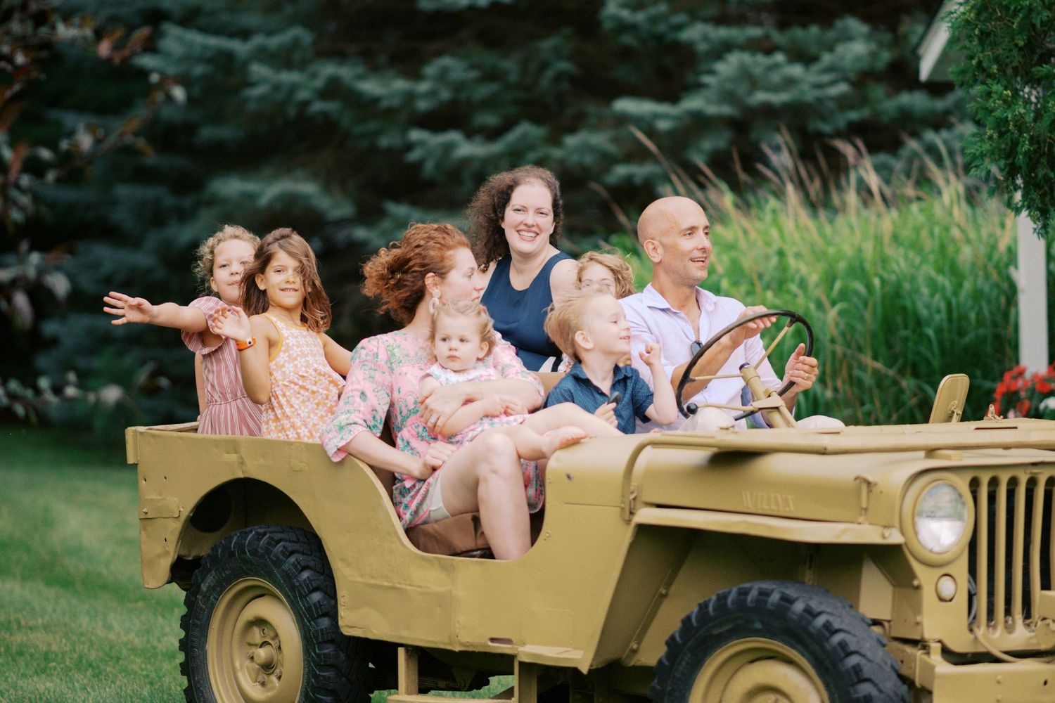 A group rides together in a vintage tan military Jeep on a sunny day with evergreen trees in the background.