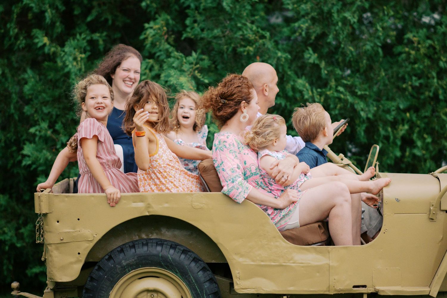 People enjoy a ride in a classic tan Jeep with lush green foliage visible behind them.
