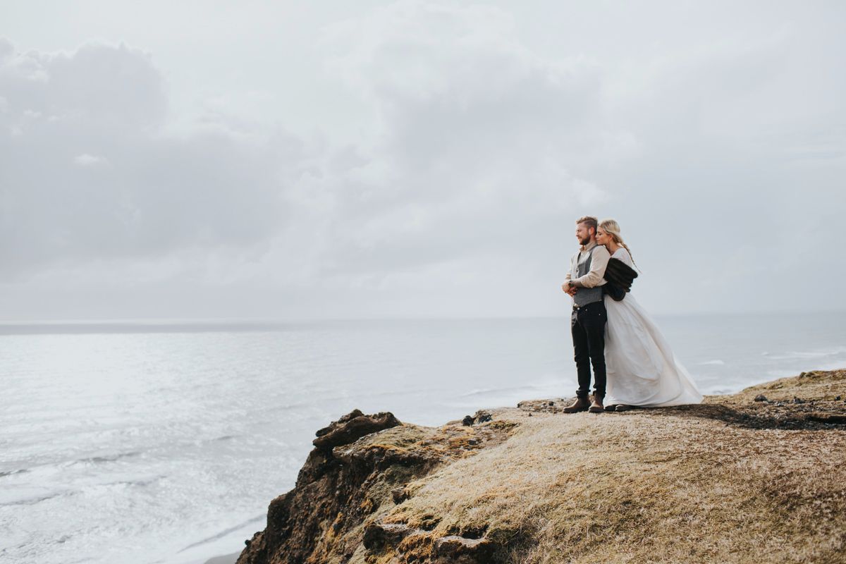 A couple in wedding attire embraces on a dramatic oceanside cliff overlooking stormy gray waters.