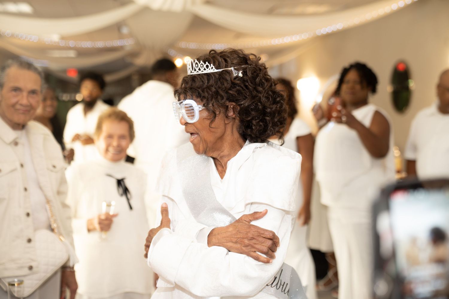 Wedding celebration scene with white-clad guests dancing and embracing under draped fabric decor.