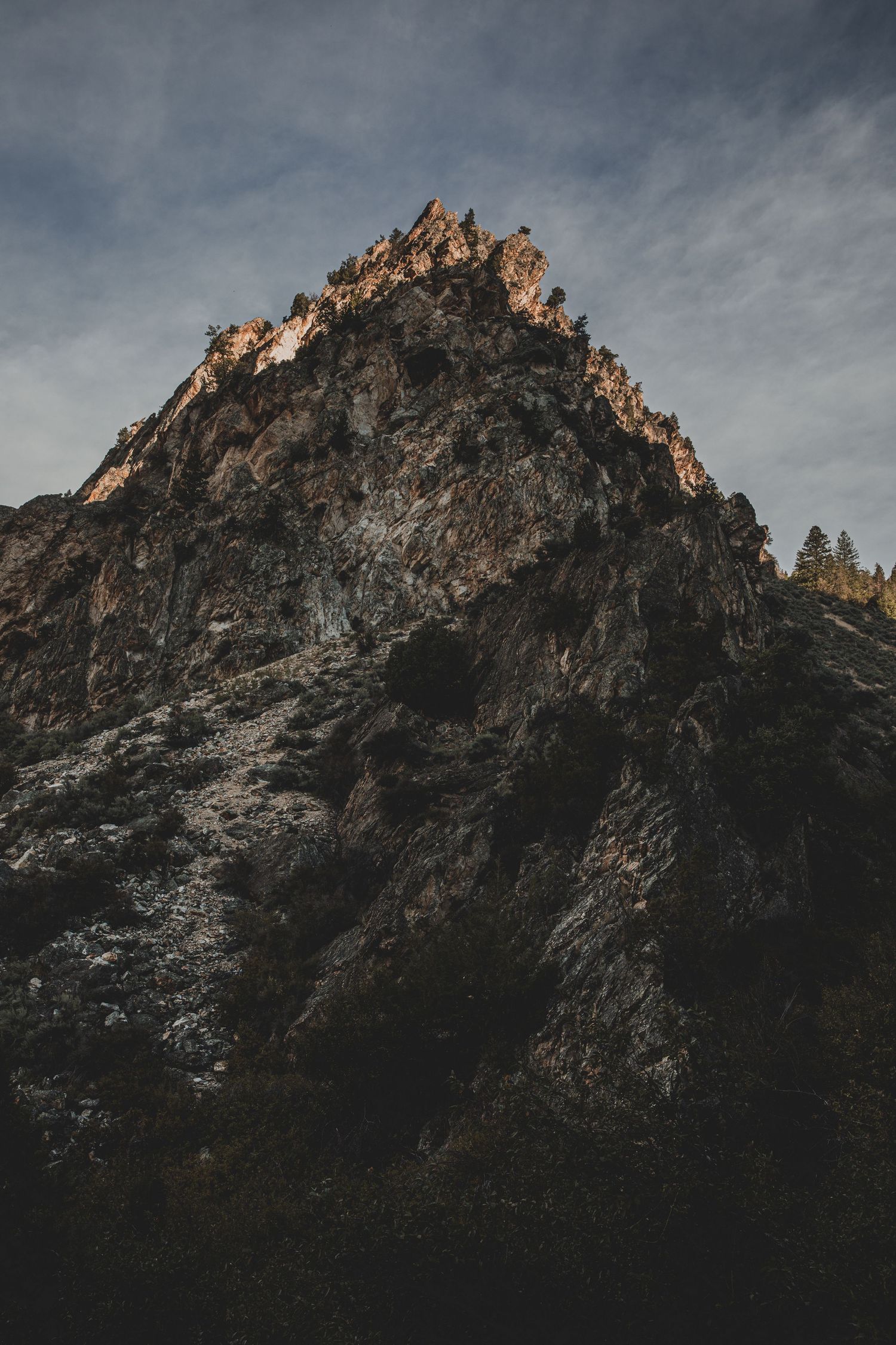 Rugged mountain peak rises dramatically against a moody sky with trees visible at its base.