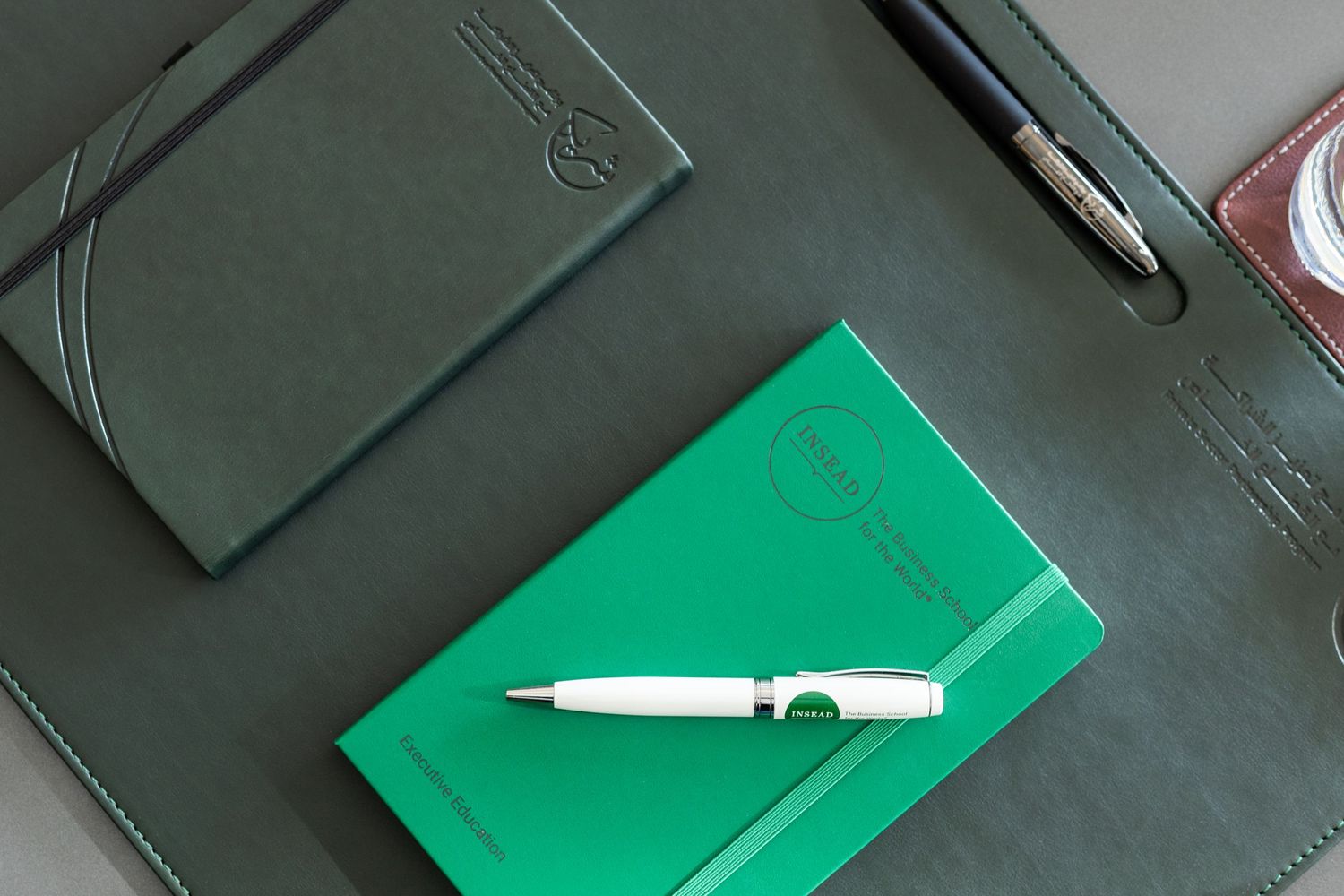 A white pen rests on a bright green notebook next to a gray leather planner on a desk surface.